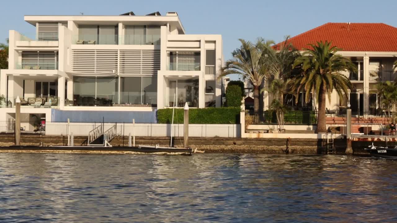 A view of contemporary riverside houses with palm trees and calm water reflecting the clear sky.