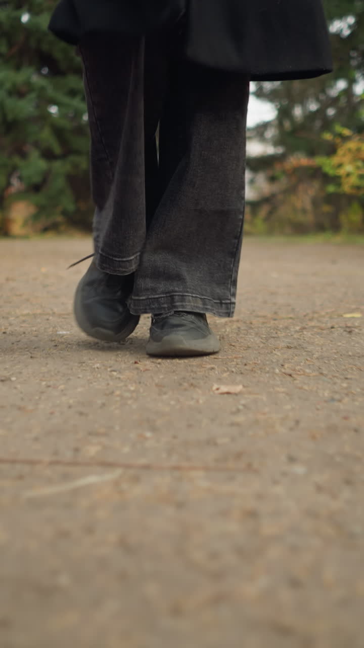 fotografía a nivel del suelo que captura las piernas de una persona con pantalones vaqueros negros y zapatos de lona caminando a través de un jardín pintoresco durante el otoño, rodeado de follaje vibrante y una atmósfera natural tranquila