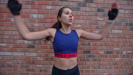 young hispanic brunette adult female in sportswear doing jumping jacks routine in an outside urban scene in front of a brick wall