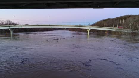 Drone-flying-under-bridge-with-cars-crossing-the-river-at-twilight-with-pink-hue-sky