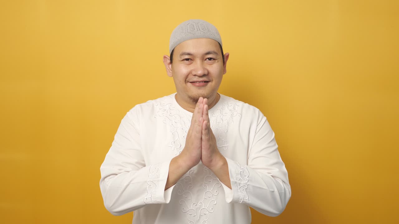 Asian muslim man smiling and shows greeting gesture against yellow background, concept of islamic celebration of ramadan or eid fitr