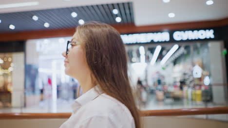 confident woman in glasses strolls through a modern shopping mall, casually glancing around, she wears a stylish white blouse and exudes elegance in a busy retail space