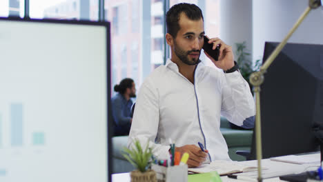 Mixed-race-businessman-sitting-at-desk-with-computer-talking-on-smartphone-in-busy-office