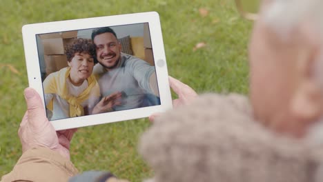 anciano sosteniendo una tableta y haciendo una videollamada de pie en el parque, una pareja saludando al otro lado de la cámara