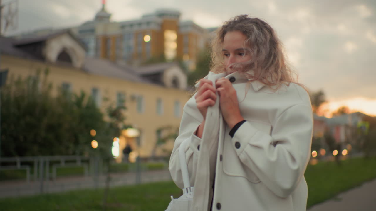 Sober cold lady covering herself with white coat while standing outdoors near residential buildings surrounded by greenery during cloudy evening with blurred background of trees and houses
