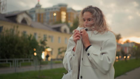 sober cold lady covering herself with white coat while standing outdoors near residential buildings surrounded by greenery during cloudy evening with blurred background of trees and houses
