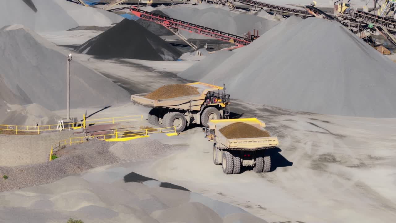 Heavy duty dump trucks loaded with gravel at quarry site, aerial tilt up to reveal conveyor belt and mounds