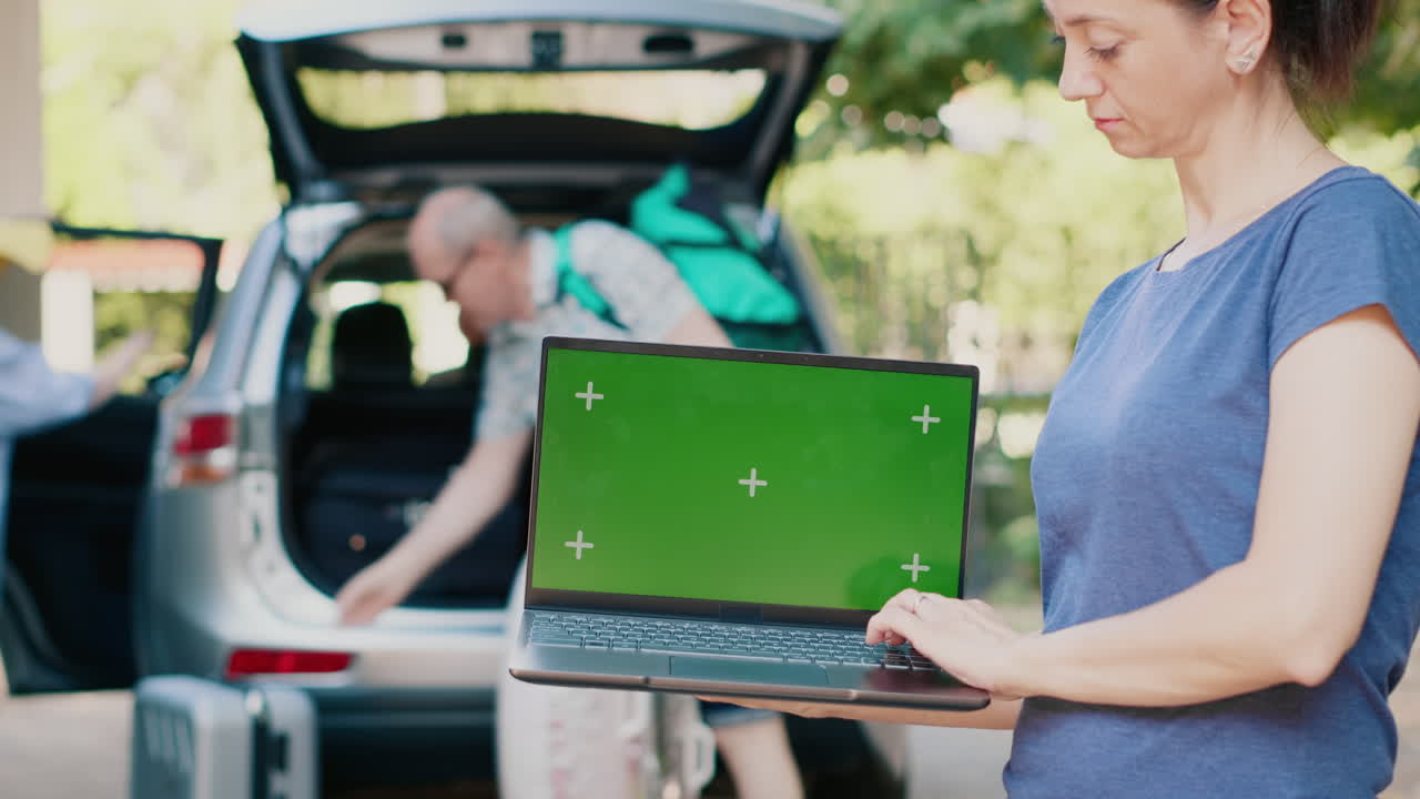 Woman using laptop with green screen while family loads car for vacation
