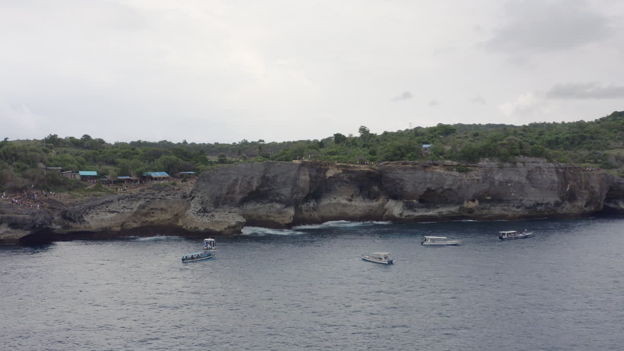 Steep high coast of Broken Beach with tourists boats drifting nearby, Nusa Penida, Bali, Indonesia. Aerial shot.