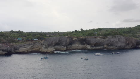 steep high coast of broken beach with tourists boats drifting nearby, nusa penida, bali, indonesia. aerial shot.