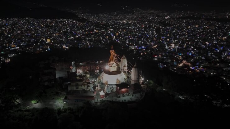 Aerial night drone footage of Swayambhunath Stupa in Kathmandu, glowing with warm lights atop the hill, showcasing its spiritual ambiance, iconic Buddhist architecture