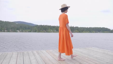 Hübsches-Mädchen-In-Leuchtend-Orangefarbenem-Kleid,-Das-Auf-Der-Holzterrasse-Am-See-Spazieren-Geht