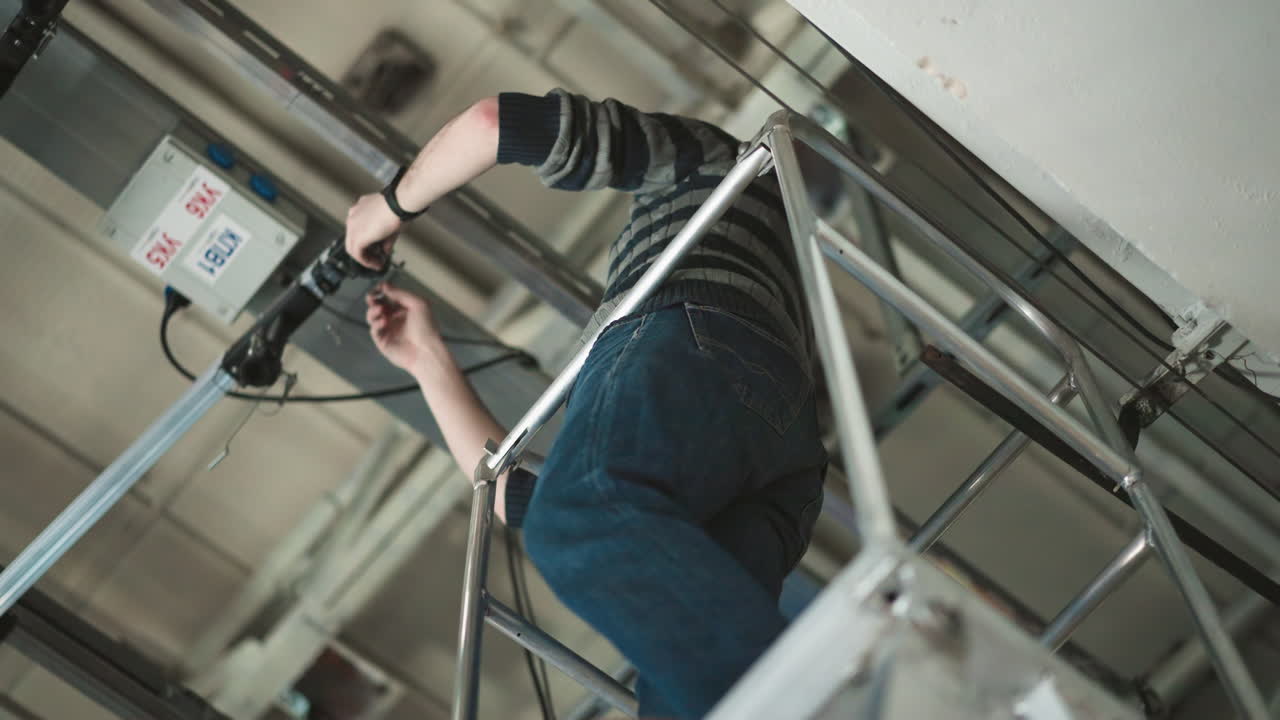 Person working at height, adjusting equipment mounted on ceiling rail system, using ladder for access, wearing sweater, gloves, fixing camera or lighting system in industrial setting