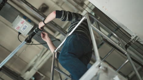 person working at height, adjusting equipment mounted on ceiling rail system, using ladder for access, wearing sweater, gloves, fixing camera or lighting system in industrial setting