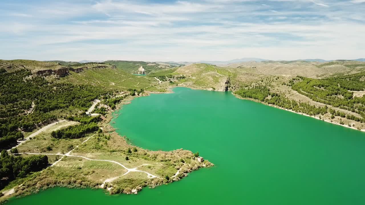 Drone footage showcasing the vibrant green waters of Embalse de la Tranquera in Spain, encircled by a stunning landscape of rolling hills, lush forests, and meandering paths under a bright blue sky