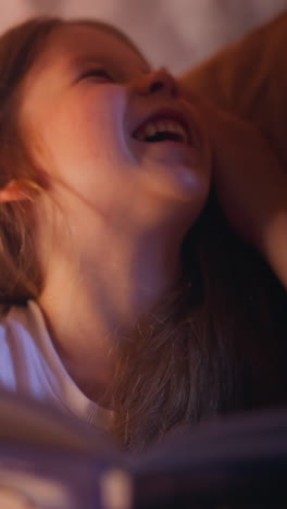 mother cuddles laughing blonde daughter holding fairy tail book on bed with pillows in semidark bedroom before going to sleep close view slow motion
