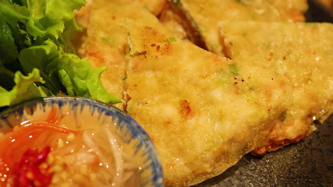 A hand reaches for a crispy shrimp pancake served with fresh greens and dipping sauce on a wooden table