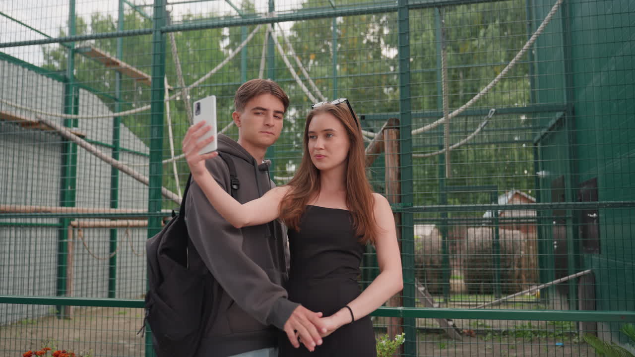 Casal caucasiano caminhando em direção ao cercado do zoológico, roupas casuais de verão, mochila, cerca de metal, corda, vasos de plantas, caminho, de mãos dadas, espontâneo, selfie com smartphone, brincalhão, carinhoso, jovem, encontro, ao ar livre