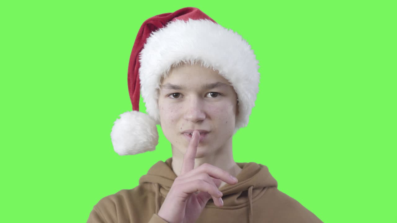 크리스마스 모자를 입은 재는 소년은 카메라에 눈을 이고 조용한 몸짓을 보여줍니다. 녹색 스크린 배경에서 포즈를 취하는 즐거운 백인 밀레니얼의 초상화.