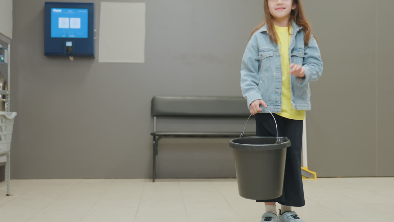 Youngster carries bucket of water, steps back from mother cleaning transparent washing machine door inside laundromat, teamwork moment, household chore, family hygiene, washers in background