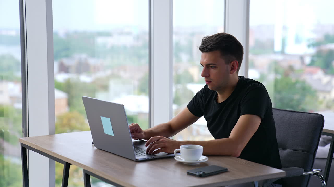 Young focused man working on laptop in office. Man takes a cup of coffee, looks at the screen and then at the window. Blurred backdrop.