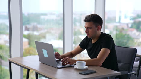 young focused man working on laptop in office. man takes a cup of coffee, looks at the screen and then at the window. blurred backdrop.