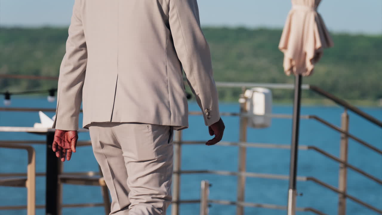 Man in beige suit walking on a scenic terrace with a view of a calm body of water