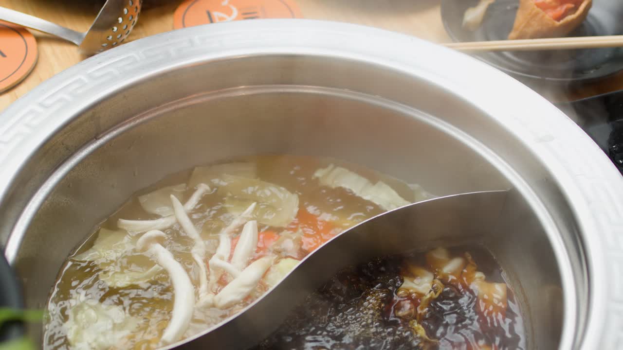 Chopsticks add vegetables and meat to steaming dual broth hotpot on a restaurant dining table