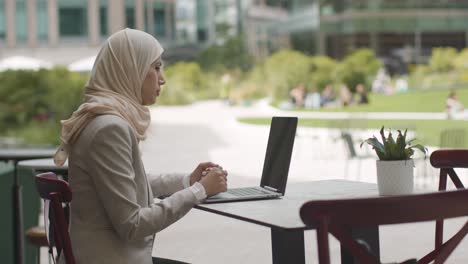 Muslim-Businesswoman-Sitting-Outdoors-In-City-Gardens-Making-Video-Call-On-Laptop
