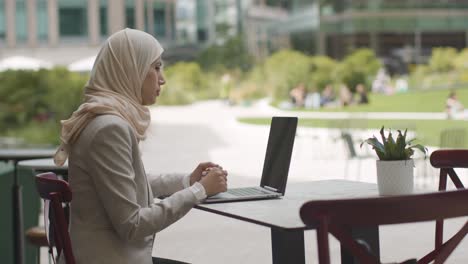 muslim businesswoman sitting outdoors in city gardens making video call on laptop