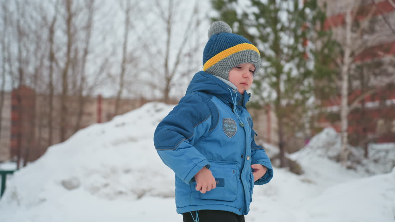 Poika heittää lumipalloa lumikasan lähellä; sinisessä takissa ja neulotussa pipossa oleva poika nostaa lunta ilmaan. Energinen liike, jalanjälkiä puiston poikki, urbaani leikkikenttä taustana. Utelias itsenäinen ja iloinen leikki