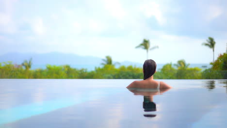 Una-Mujer-De-Espaldas-A-La-Cámara-Mira-Desde-La-Piscina-De-Un-Resort-Al-Paraíso-Tropical-Y-Al-Océano-Más-Allá