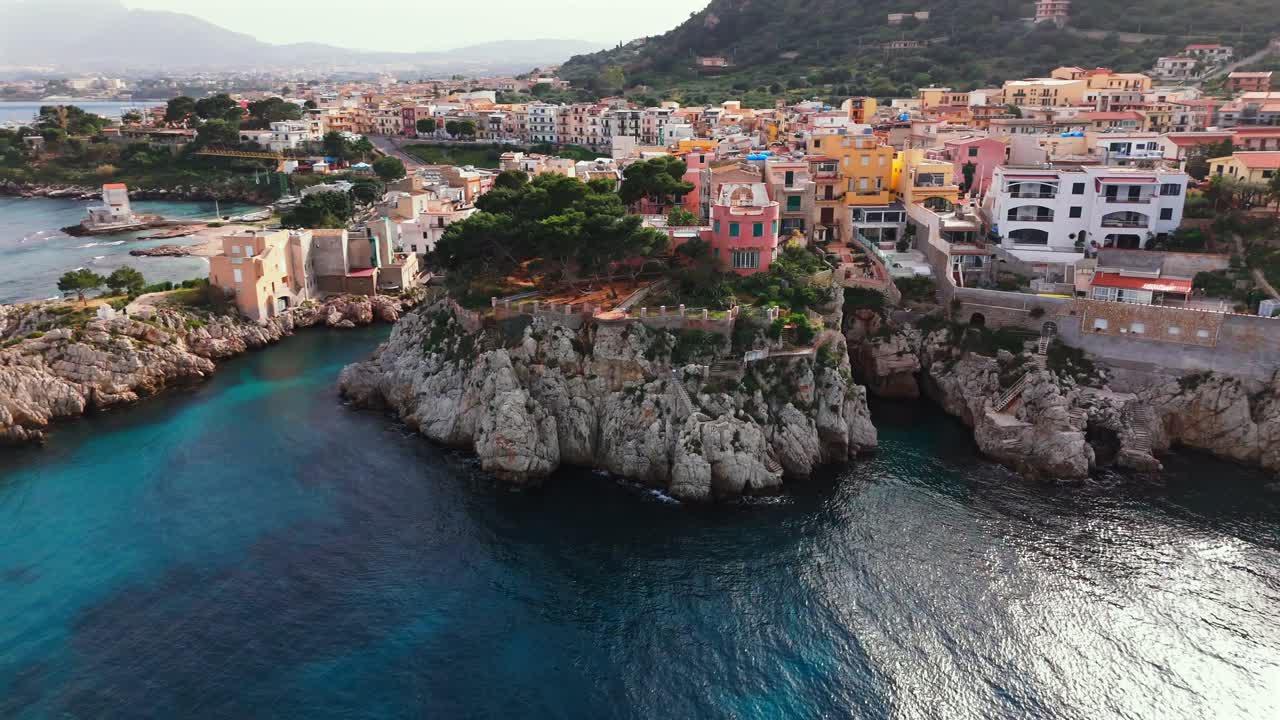 Drone flies sideways along the Porticello coastline, passing a pink cliffside villa, lush trees, rocky shore and colorful Mediterranean buildings near Palermo in northern Sicily