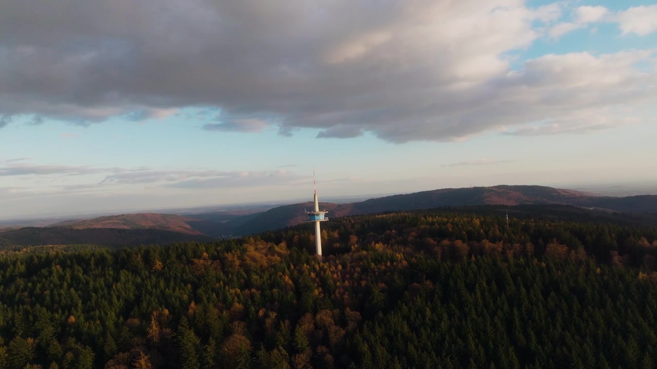 Oblet dronu kolem věže nad lesem Odenwald, odhalující podzimní lesní krajinu, zvlněné kopce a dramatické mraky v teplém večerním světle.