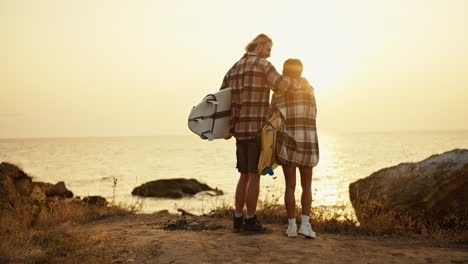 Rear-view-of-a-tall-blond-guy-in-a-plaid-shirt-and-shorts-and-his-blond-girlfriend-in-a-hat-and-plaid-shirt-holding-surfboards-in-their-hands-and-standing-on-the-earthen-seashore-looking-at-the-sunrise-in-the-morning-in-summer