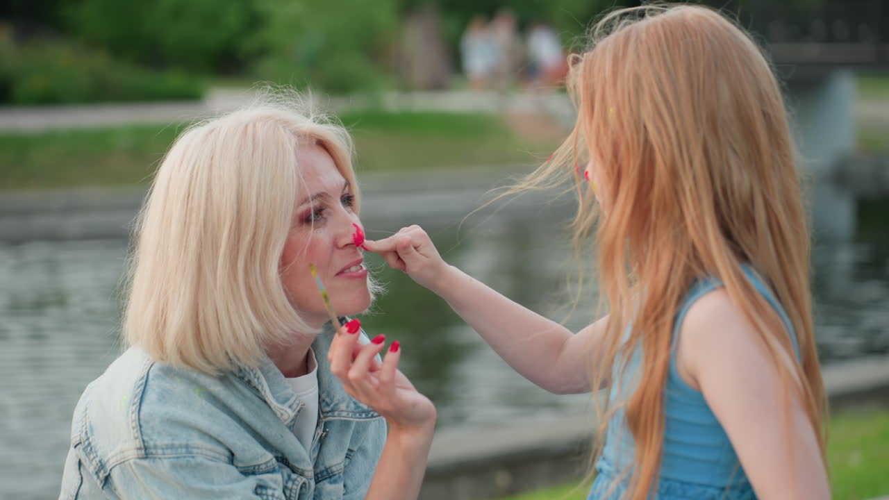 elegant mother in denim jacket sitting by calm riverbank at golden hour while daughter gently massages bright paint on her nose with little fingers in playful outdoor art moment under evening glow