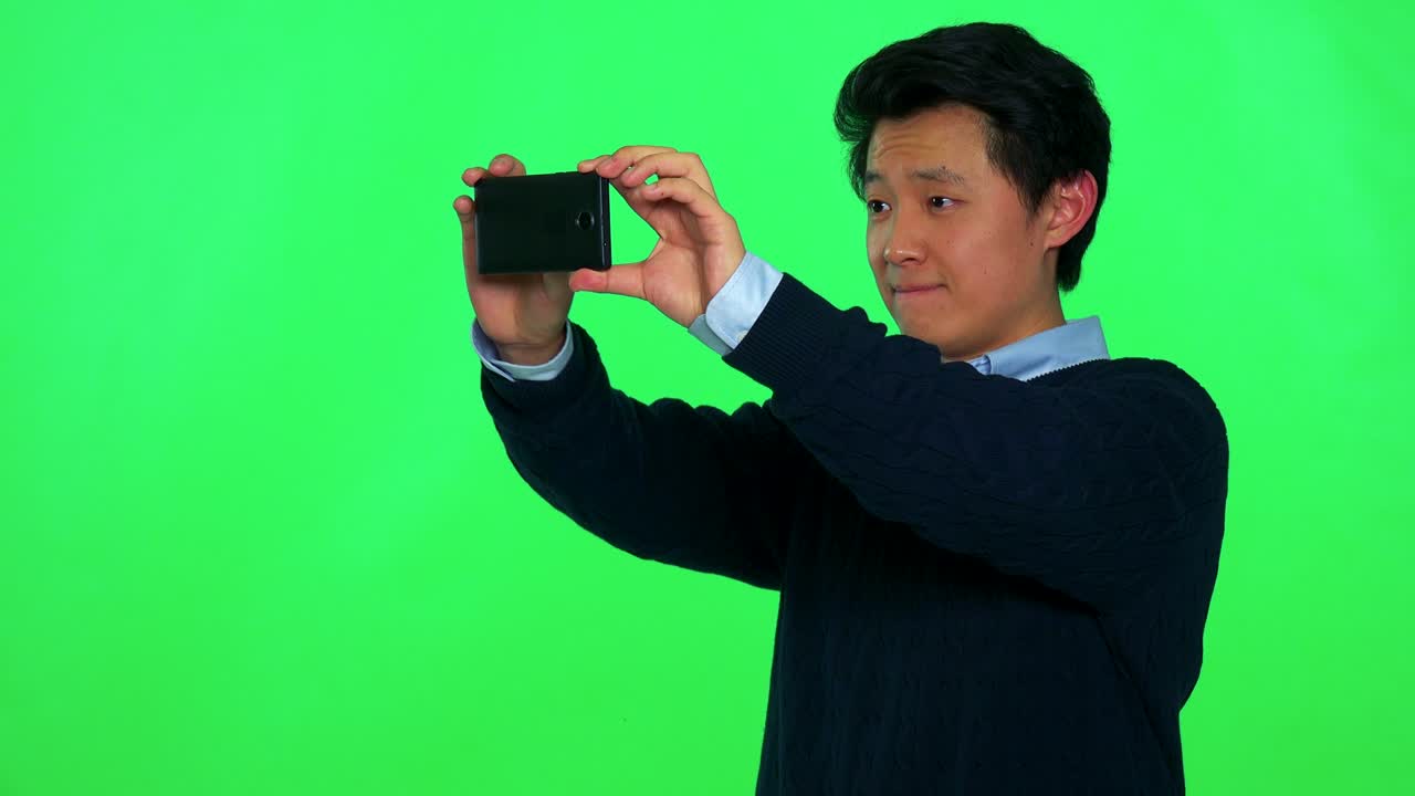젊은 아시아 남자가 스마트폰으로 카메라에서 무언가를 찍습니다. - 그린 스크린 스튜디오