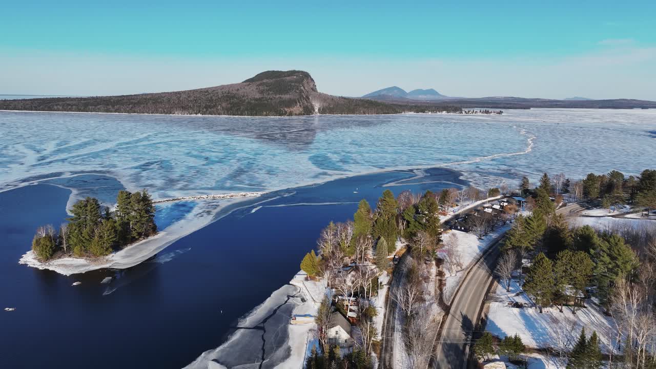 Aerial view of Rockwood, Maine in winter, showcasing a partially frozen Moosehead Lake with Mount Kineo in the background. The landscape features snowy roads, evergreen trees, and scattered cabins.