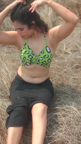 woman in green top tying hair while sitting on dry grass under bright sunlight