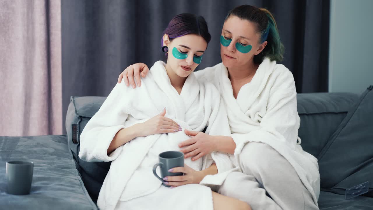 Pregnant lesbian woman and her partner are happy bonding on sofa together in bathrobes and patches