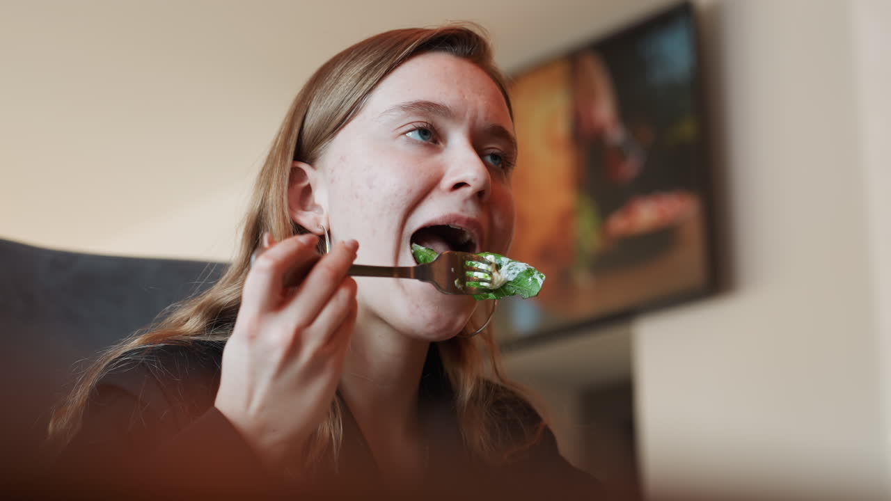 Mujer joven probando ensalada de hojas verdes en interiores, con un primer plano de su expresión vacilante. Un tenedor con verduras, un fondo de sala de estar, iluminación cálida y suave, y un suéter casual, capturan un momento cotidiano espontáneo de sutil decepción.