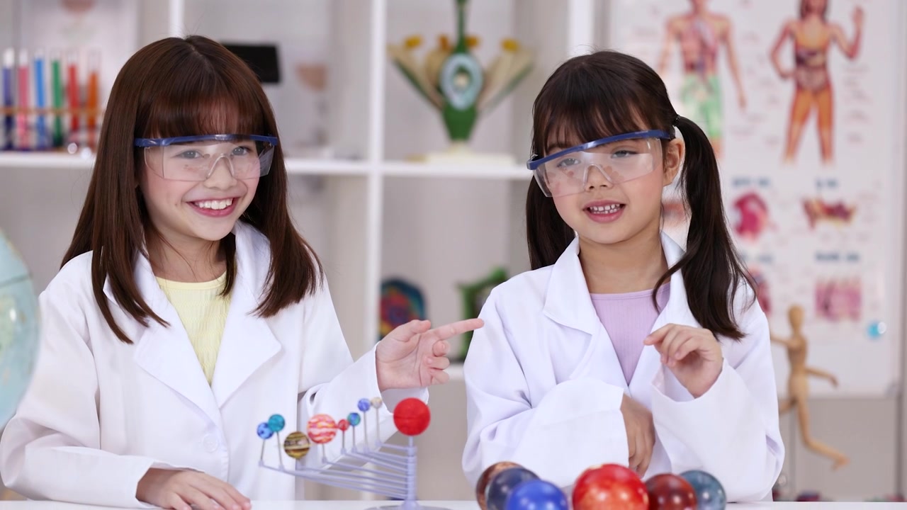 Kaksi koulutyttöä, joilla on laboratoriotakit ja suojalasit, tutkivat planeettamallia luonnontiedon tunnilla.