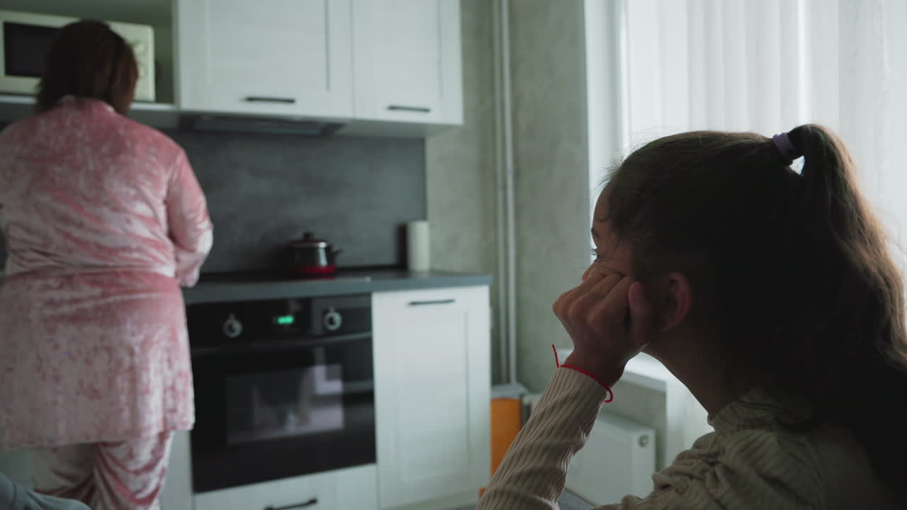 Young girl with ponytail thoughtfully watches woman in pink velvet robe preparing breakfast in modern kitchen, sunlight shining through curtain-covered window, capturing quiet family morning moment indoors