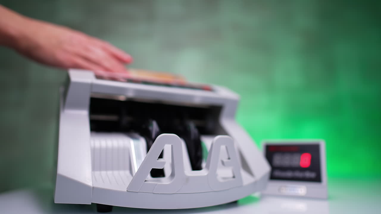Hands places money into the cash counting machine. Bills counted in equipment fall on the holder. Blurred backdrop.