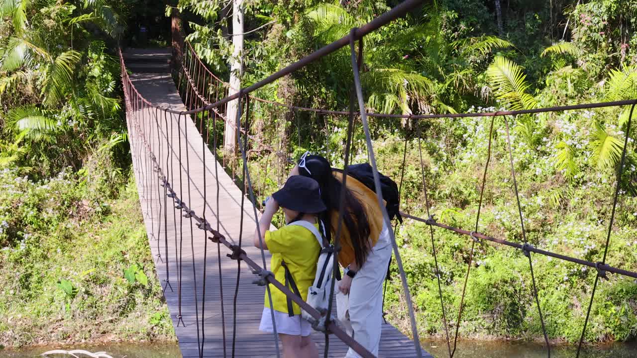 Uma mãe e uma criança estão em uma ponte suspensa de madeira em uma floresta tropical exuberante