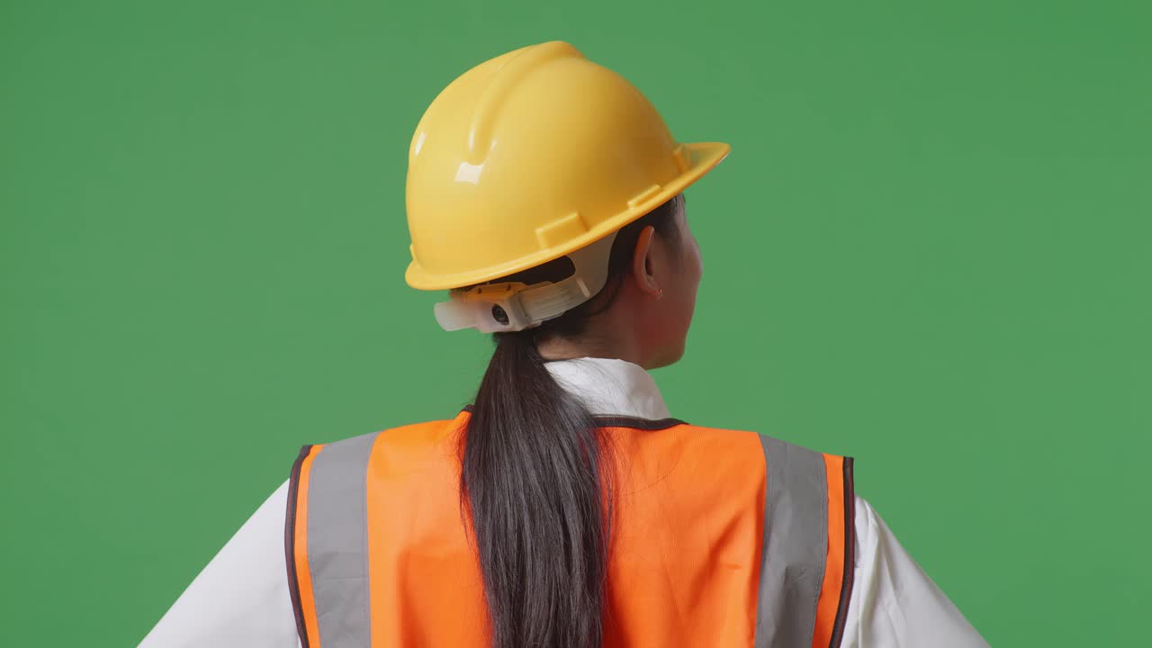 안전 헬을 착용 한 여성 엔지니어가 녹색 스크린 배경 스튜디오에서 팔을 들고 서있는 동안 주위를 둘러보고 있는 근접한 뒷면