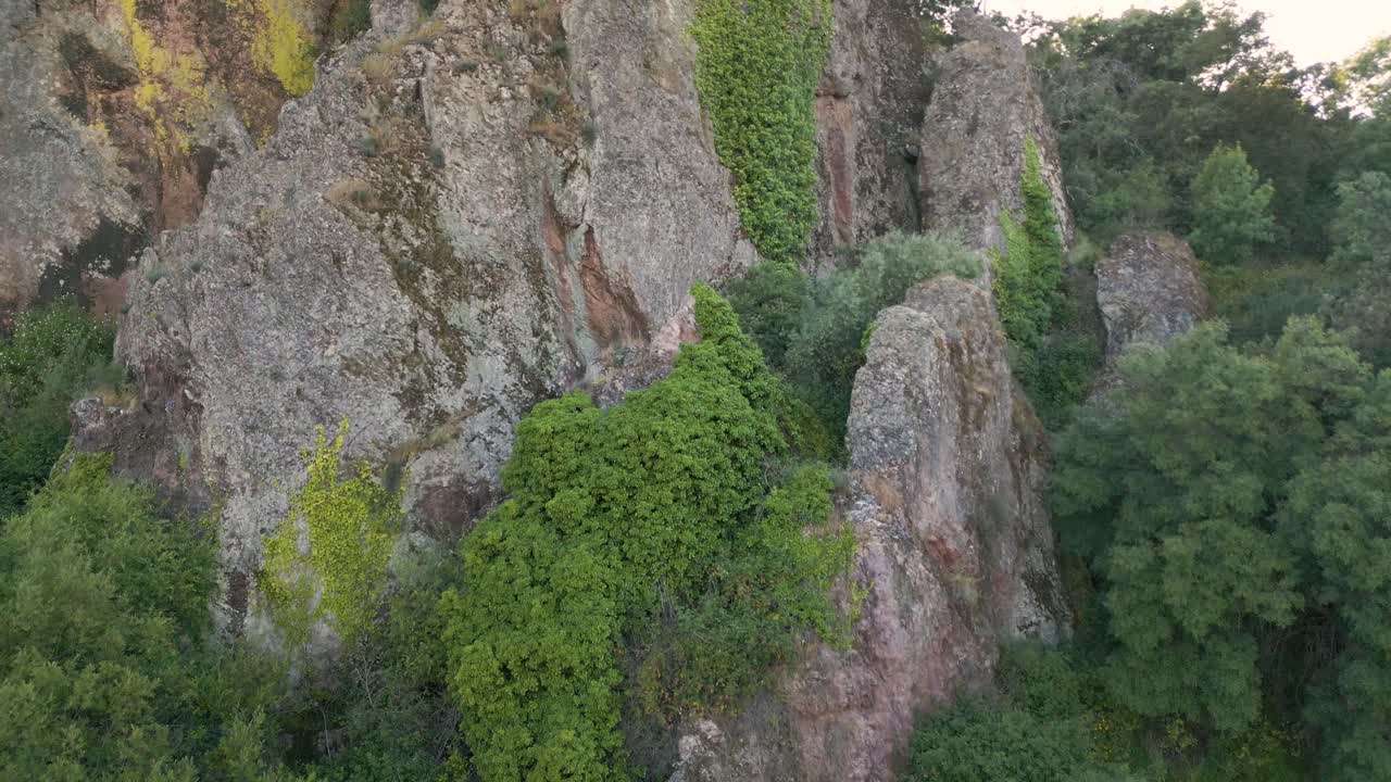 Aerial drone shot approaching limestone rock formation in Villarta de los Montes, Badajoz. The stones glow in gray, orange, and green tones under sunlight, surrounded by bright green trees and vines