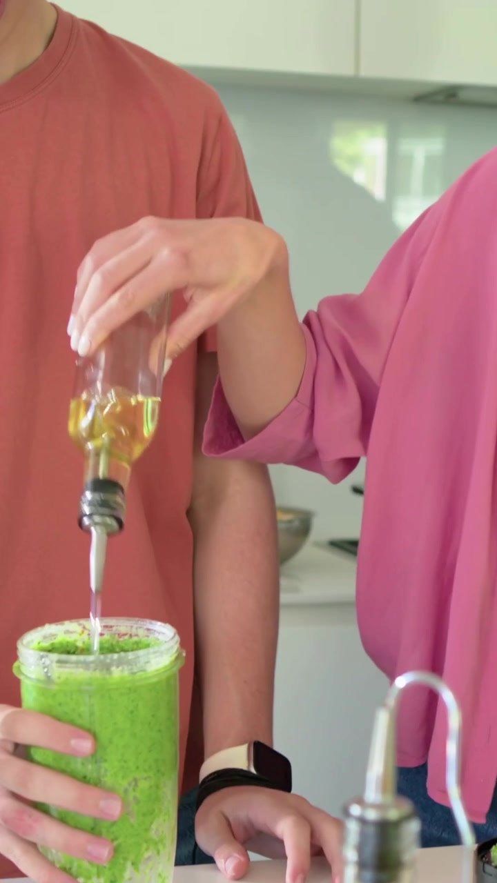 Video vertical: Mujer con top rosa tomando aceite, vertiéndolo en licuadora en el mostrador para hacer una bebida verde.