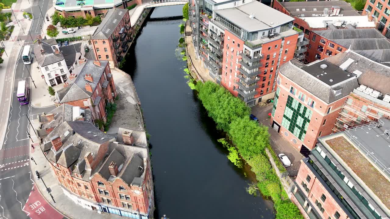 Drone footage captures a turquoise bus traveling across a city bridge above the River Aire, surrounded by Victorian brick buildings in Leeds, England. Daylight, smooth camera movement
