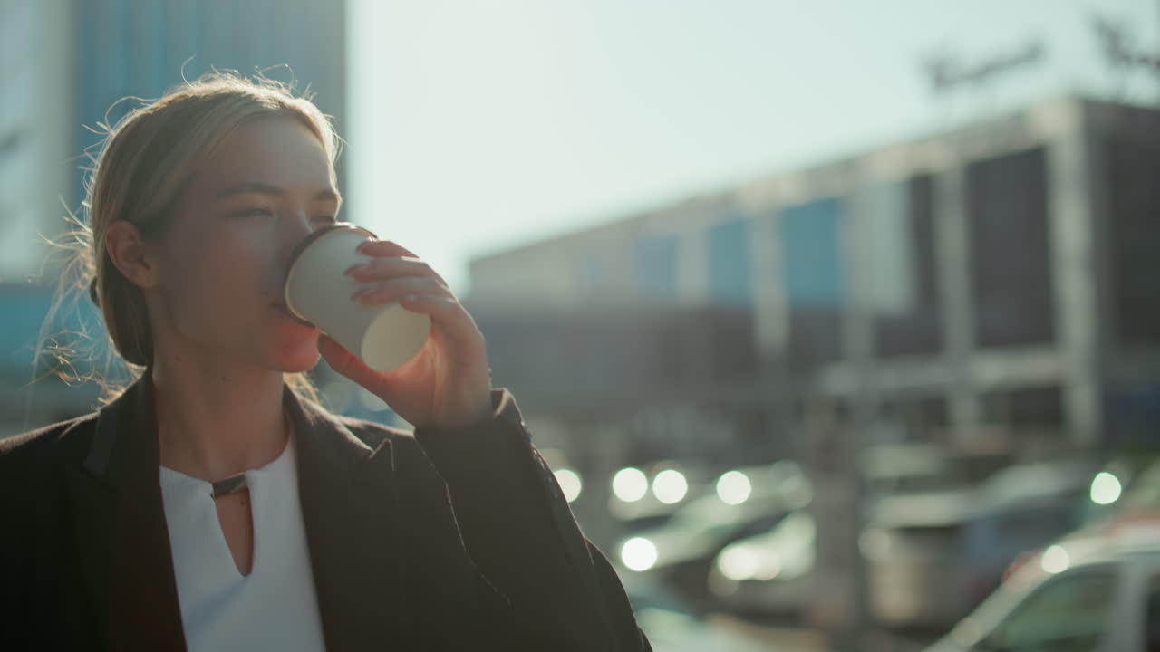Confident lady bathed in warm glow walks while drinking coffee, wearing professional attire with blurred background of residential building, parked cars, and soft sunlight creating a radiant ambiance